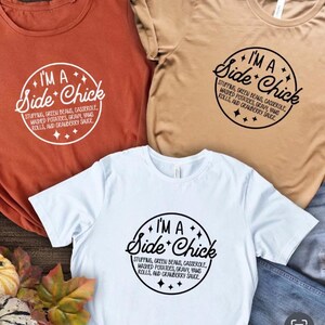 May include: Three t-shirts in rust, tan, and white, each with the phrase "I'M A Side Chick" in a circular design. The design includes the words "Stuffing, Green Beans, Casserole, Mashed Potatoes, Gravy, Yams, Rolls, and Cranberry Sauce."