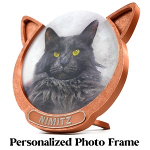 Puede incluir: Un marco de fotos en forma de gato de color cobre con una foto de un gato negro con ojos amarillos. El marco tiene un soporte y el nombre "NIMITZ" está grabado en la parte inferior. El texto "Personalized Photo Frame" está debajo.