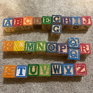 May include: A collection of wooden alphabet blocks arranged in three rows. Each block features a letter of the alphabet in a primary color on one side and a corresponding black line drawing on the other. The letters A through Z are present.