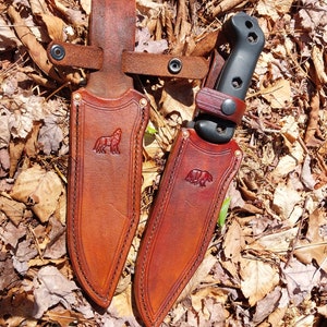 Puede incluir: Dos fundas de cuero con un diseño de lobo y oso, para cuchillos con mangos negros. Las fundas son de color marrón con un borde cosido y están sobre un lecho de hojas marrones.