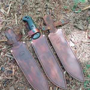 Puede incluir: Tres cuchillos con fundas de cuero marrón. Las fundas tienen un emblema de oso y están cosidas con hilo negro. Un cuchillo tiene un mango negro y una correa de cuero. Los cuchillos están sobre un lecho de hojas y ramitas.