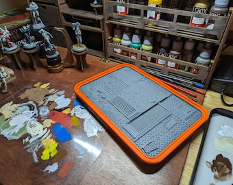 Puede incluir: Una paleta de pintura de plástico gris texturizado con un borde naranja. La paleta tiene una variedad de texturas y patrones, incluyendo ladrillo, panal de abeja y placa de diamante. La paleta está sobre una mesa de madera con una variedad de colores de pintura y figuras en miniatura.