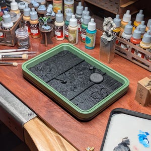 Puede incluir: Una bandeja de plástico verde con una superficie negra texturizada, utilizada para pintar miniaturas. La bandeja está sobre una mesa de madera con una variedad de botellas de pintura y figuras en miniatura.