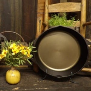 Könnte beinhalten: Eine schwarze gusseiserne Bratpfanne mit einem Ständer, die auf einer Holzoberfläche steht. Die Bratpfanne ist leer und hat einen glänzenden, goldfarbenen Innenraum. Ein Strauß gelber Blumen steht in einer Vase links neben der Bratpfanne.