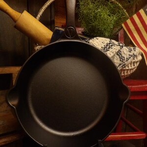 May include: A black cast iron skillet with a long handle and a helper handle. The skillet has a dark, textured surface and is displayed on a black stand. The handle has a hole for hanging. A rolling pin and a basket are in the background.