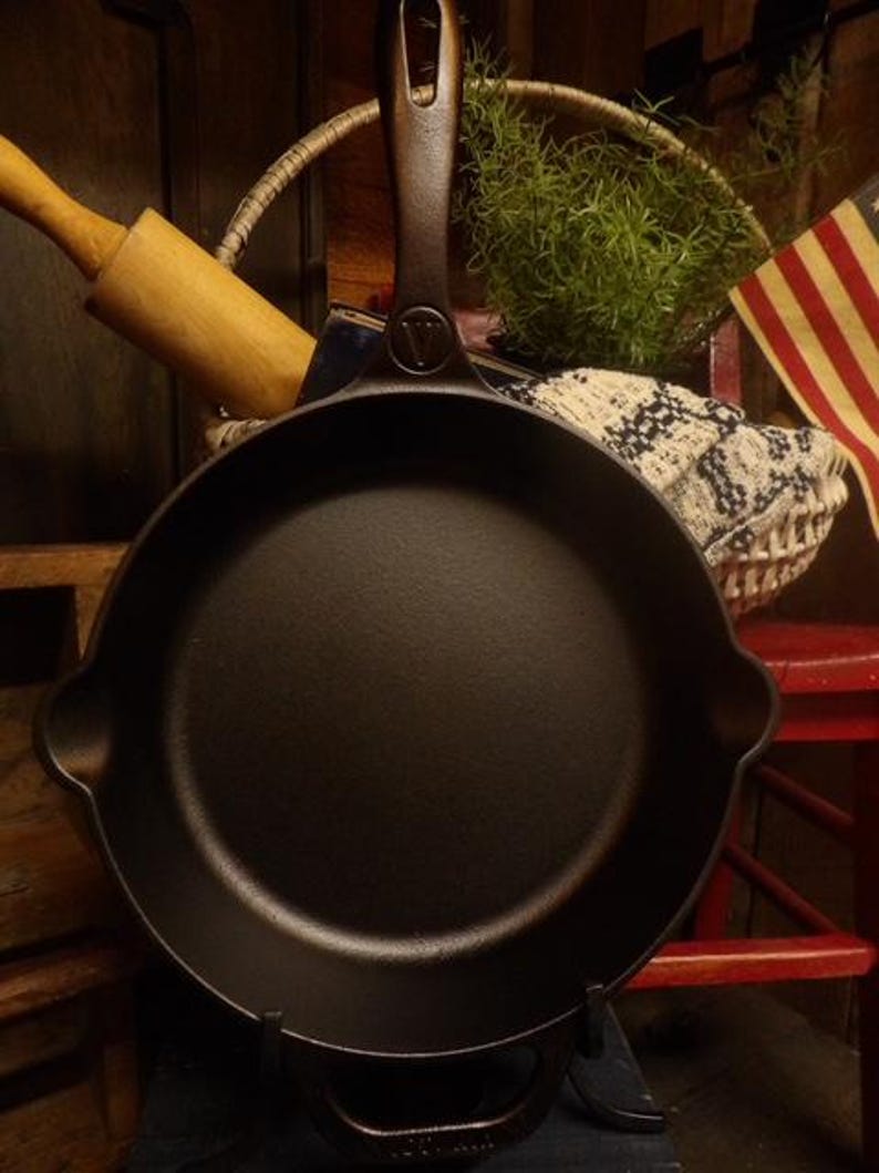 May include: A black cast iron skillet with a long handle and a helper handle. The skillet has a dark, textured surface and is displayed on a black stand. The handle has a hole for hanging. A rolling pin and a basket are in the background.