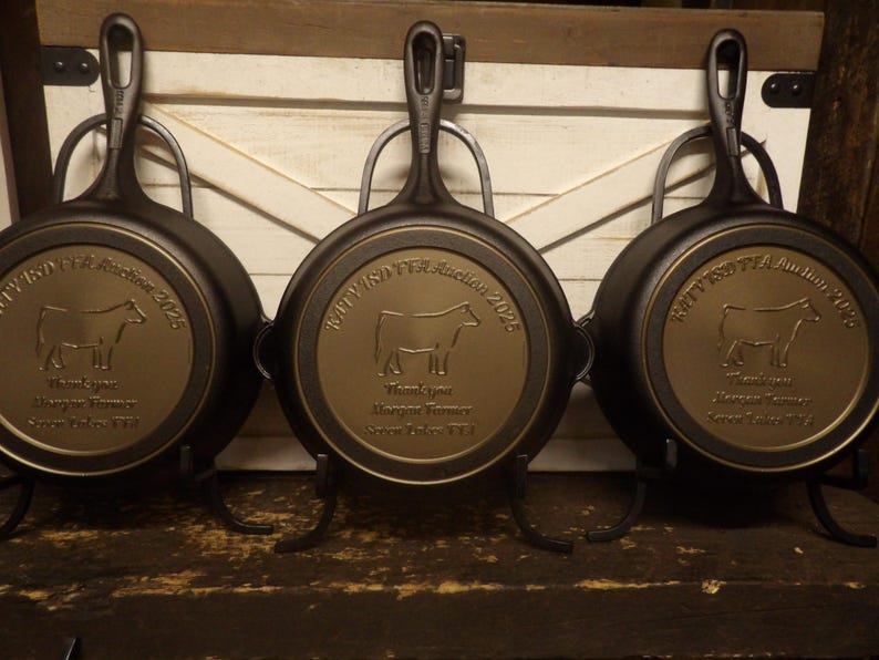 May include: Three cast iron skillets with a bronze-colored medallion featuring a cow silhouette and text. The skillets are displayed on black metal stands against a wooden background. The text includes "BTV FFA Auction 2025".