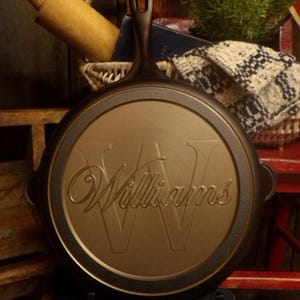 May include: A cast iron skillet with a handle and the name "Williams" embossed on the cooking surface. The skillet is dark brown with a gold-colored cooking surface. The skillet is sitting on a black stand.