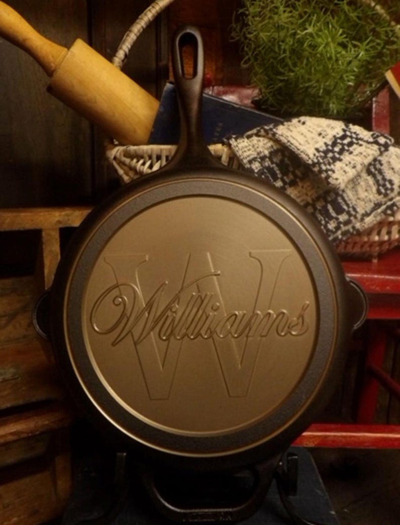 May include: A cast iron skillet with a handle and the name "Williams" embossed on the cooking surface. The skillet is dark brown with a gold-colored cooking surface. The skillet is sitting on a black stand.