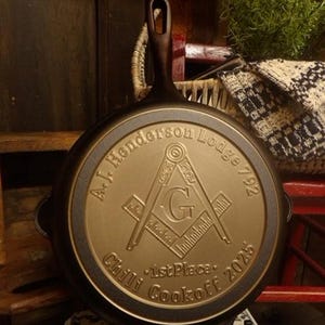 May include: A cast iron skillet with a handle and a decorative emblem. The emblem features the text "A.J. Henderson Lodge 792" and "Chili Cookoff 2025". The skillet is displayed on a black metal stand.