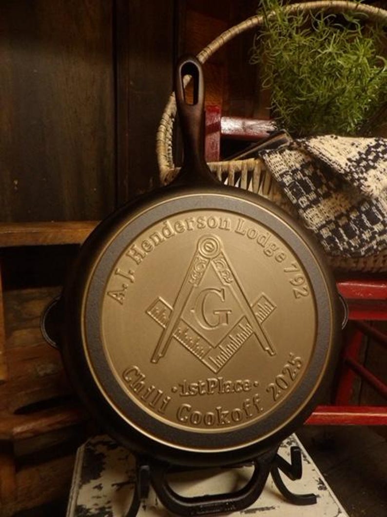 May include: A cast iron skillet with a handle and a decorative emblem. The emblem features the text "A.J. Henderson Lodge 792" and "Chili Cookoff 2025". The skillet is displayed on a black metal stand.