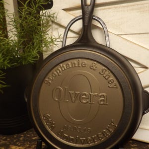 May include: A black cast iron skillet with a long handle, resting on a black metal stand. The skillet's underside features the engraved names "Stephanie & Shey Olvera" and the date "06.03.19" commemorating a 6th anniversary.