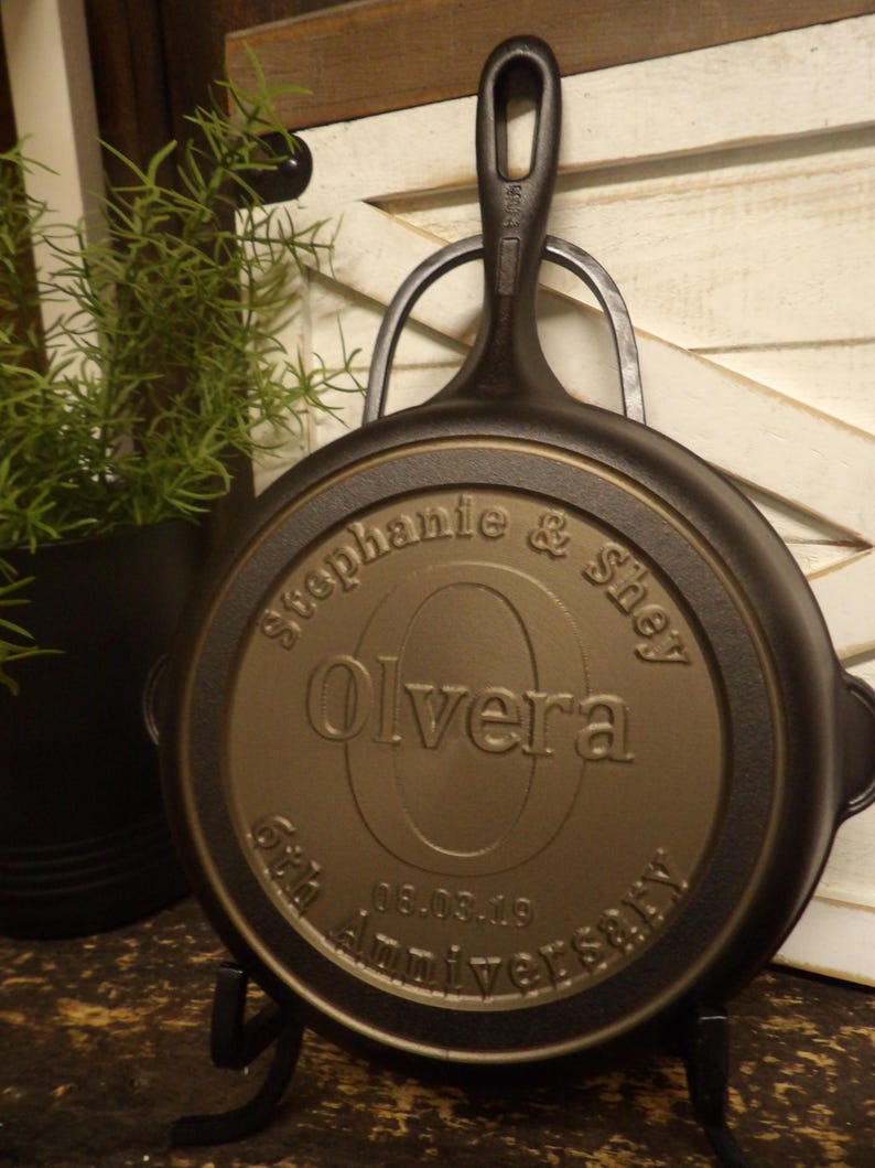 May include: A black cast iron skillet with a long handle, resting on a black metal stand. The skillet's underside features the engraved names "Stephanie & Shey Olvera" and the date "06.03.19" commemorating a 6th anniversary.