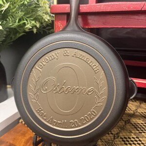 May include: A black cast iron skillet with a bronze-colored center. The center is engraved with the names "Jeremy & Amanda" above the name "Osborne" and the date "Est. April 20, 2020". The skillet is on a black metal stand.