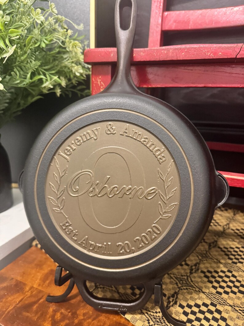 May include: A black cast iron skillet with a bronze-colored center. The center is engraved with the names "Jeremy & Amanda" above the name "Osborne" and the date "Est. April 20, 2020". The skillet is on a black metal stand.
