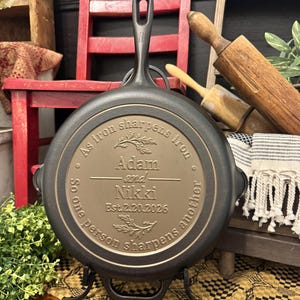 May include: A black cast iron skillet with a long handle and a decorative inscription. The inscription reads "As iron sharpens iron, so one person sharpens another" with the names "Adam and Nikki" and the date "Est.2.20.2026".
