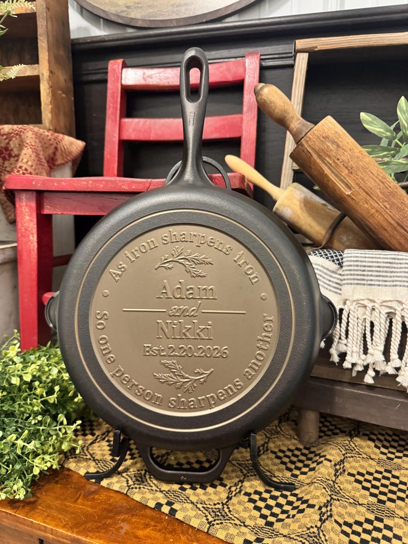 May include: A black cast iron skillet with a long handle and a decorative inscription. The inscription reads "As iron sharpens iron, so one person sharpens another" with the names "Adam and Nikki" and the date "Est.2.20.2026".