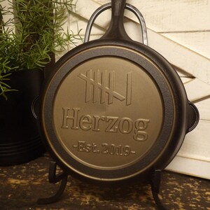 May include: A black cast iron skillet with a gold-colored center featuring the name "Herzog" and "Est. 2019". The skillet is displayed on a black metal stand, with a plant in a black pot in the background.