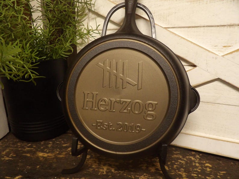 May include: A black cast iron skillet with a gold-colored center featuring the name "Herzog" and "Est. 2019". The skillet is displayed on a black metal stand, with a plant in a black pot in the background.