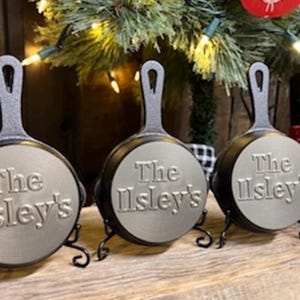 May include: Three cast iron skillet ornaments with the text "The Ilsley's" embossed on the front. Each skillet is black with a silver-colored cooking surface and a handle. They are displayed on decorative black metal stands.