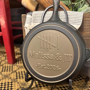 May include: A black cast iron skillet with a handle, resting on a decorative stand. The skillet's interior features the engraved names "Melissa & JT" and the year "Est. 2026". The skillet is displayed on a yellow and black patterned surface.