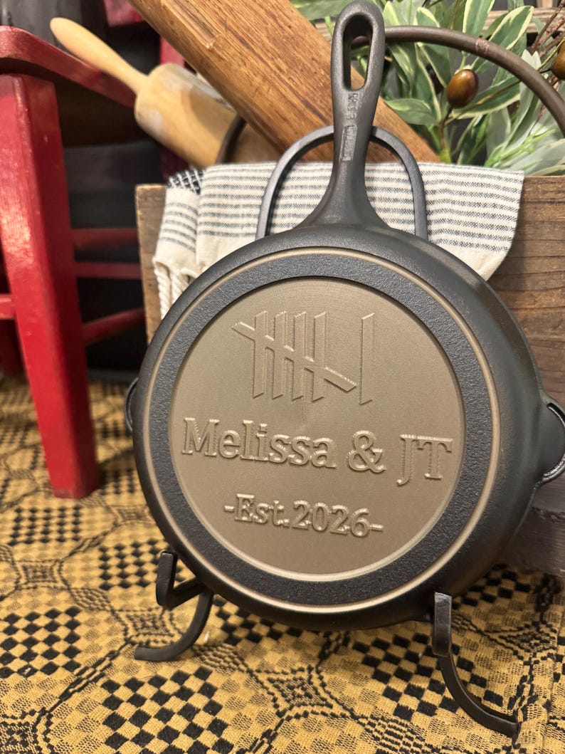 May include: A black cast iron skillet with a handle, resting on a decorative stand. The skillet's interior features the engraved names "Melissa & JT" and the year "Est. 2026". The skillet is displayed on a yellow and black patterned surface.