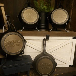 May include: Several black cast iron skillets with the text "JAMESON BLACK BARREL" on the bottom. Some skillets are displayed on stands. The background includes a white wooden chest and a small plant.