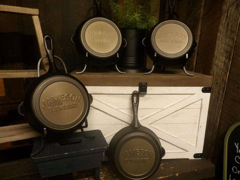 May include: Several black cast iron skillets with the text "JAMESON BLACK BARREL" on the bottom. Some skillets are displayed on stands. The background includes a white wooden chest and a small plant.