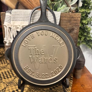 May include: A black cast iron skillet with the words "LOVE YOU MORE!" and "The Wards EST. 3.22.2020" engraved on the bottom. The skillet is displayed on a black metal stand. A rolling pin and a striped towel are in the background.