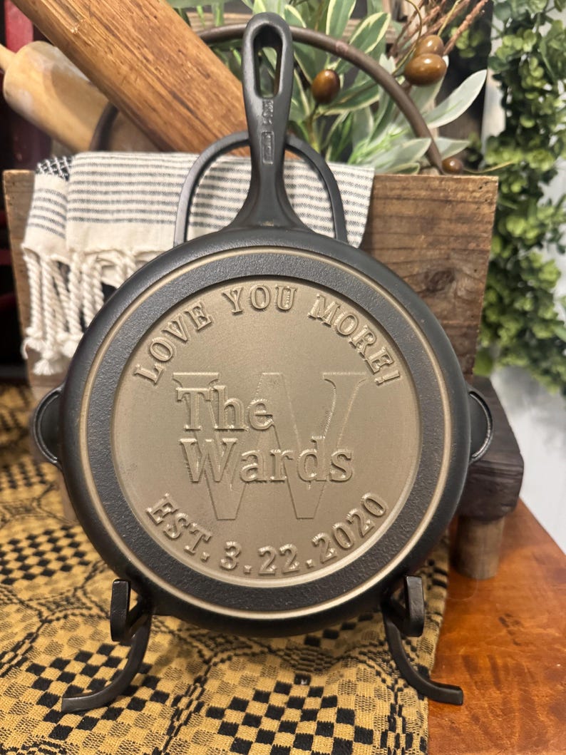 May include: A black cast iron skillet with the words "LOVE YOU MORE!" and "The Wards EST. 3.22.2020" engraved on the bottom. The skillet is displayed on a black metal stand. A rolling pin and a striped towel are in the background.
