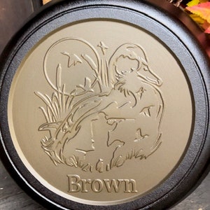 May include: A black cast iron skillet with a brown, embossed design of a duck and reeds. The skillet has the word "Brown" inscribed on the bottom.