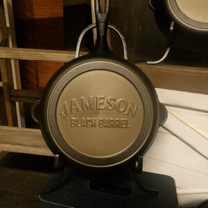May include: A black cast iron skillet with the words "JAMESON BLACK BARREL" embossed on the bottom. The skillet is displayed on a black metal stand. The handle has a hole for hanging. Another skillet is visible in the background.