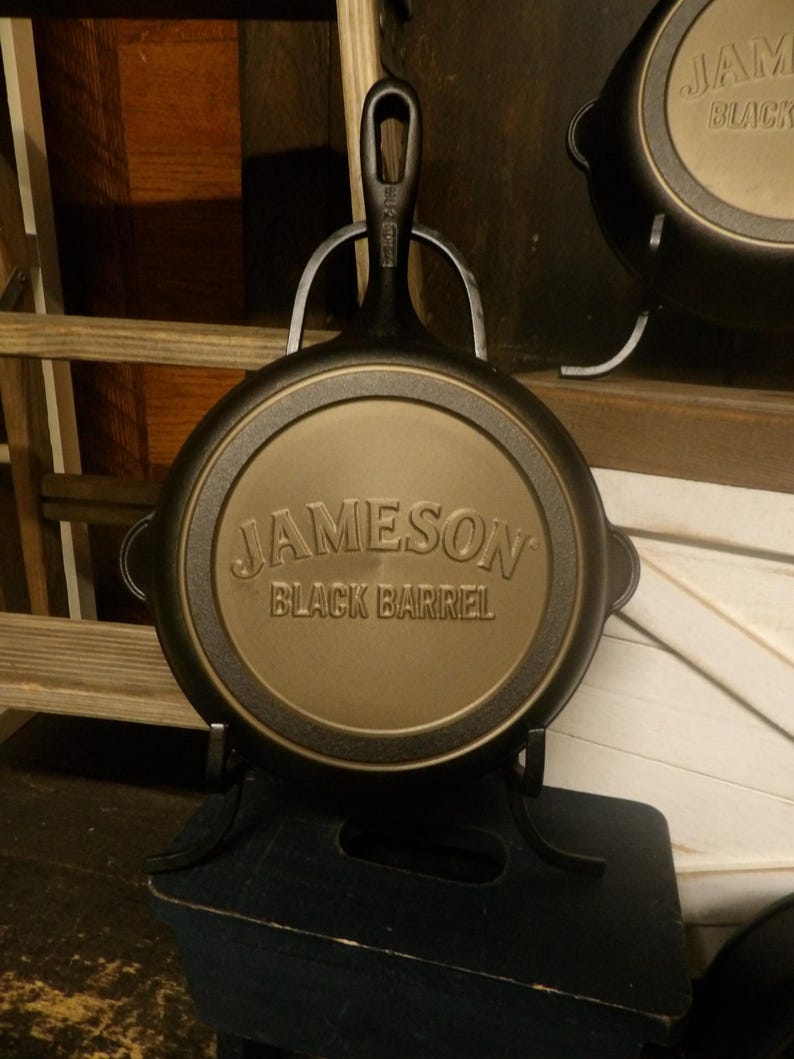 May include: A black cast iron skillet with the words "JAMESON BLACK BARREL" embossed on the bottom. The skillet is displayed on a black metal stand. The handle has a hole for hanging. Another skillet is visible in the background.