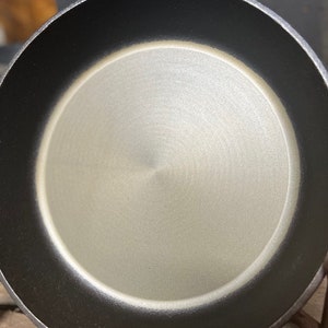May include: A black cast iron skillet with a smooth, light gray interior. The skillet has a handle and is sitting on a wooden surface.