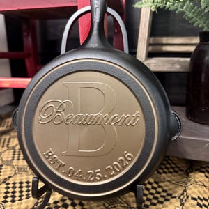May include: A black cast iron skillet with a bronze-colored center. The center features the name "Beaumont" above a large letter "B" and the date "EST. 04.25.2026". The skillet is displayed on a black metal stand.