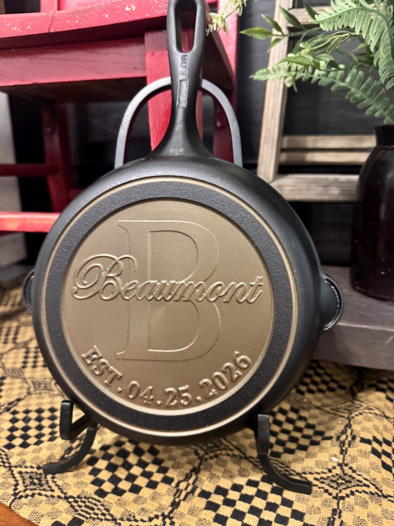 May include: A black cast iron skillet with a bronze-colored center. The center features the name "Beaumont" above a large letter "B" and the date "EST. 04.25.2026". The skillet is displayed on a black metal stand.