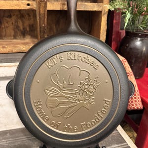May include: A black cast iron skillet with a long handle, resting on a black metal stand. The skillet features a circular design with the text "KT's Kitchen Home of the Foodfeed" and a chef's hat, utensils, and floral elements.
