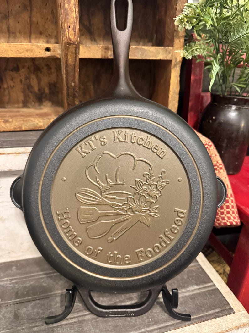May include: A black cast iron skillet with a long handle, resting on a black metal stand. The skillet features a circular design with the text "KT's Kitchen Home of the Foodfeed" and a chef's hat, utensils, and floral elements.