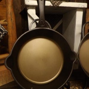 Może przedstawiać: Czarna patelnia żeliwna z długim uchwytem i gładkim, złotobrązowym wnętrzem. Patelnia stoi na czarnym metalowym stojaku.