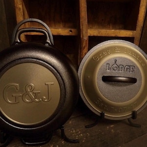 May include: Two cast iron cookware pieces. One is a round, black pot with a handle and a gold-colored circle with the letters "G&J". The other is a lid with the text "Greg & Janice" and "Lodge". Both pieces are on black stands.
