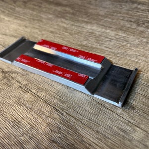 May include: A silver metal strip with two strips of red 3M adhesive tape on the top and bottom. The strip has a channel in the middle.