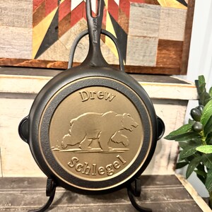 May include: A black cast iron skillet with a gold-colored medallion featuring a bear and the text "Drew Schlegel." The skillet has a handle with a hole for hanging and is displayed on a black metal stand.