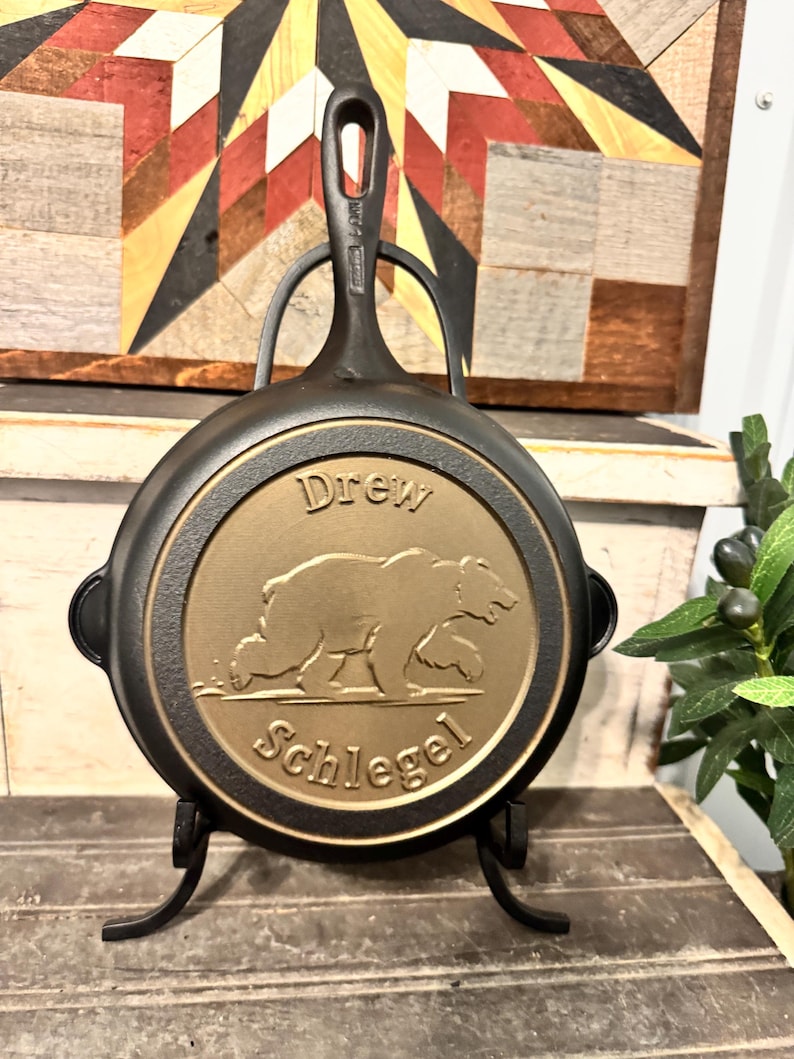 May include: A black cast iron skillet with a gold-colored medallion featuring a bear and the text "Drew Schlegel." The skillet has a handle with a hole for hanging and is displayed on a black metal stand.