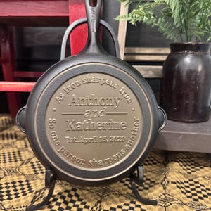 May include: A cast iron skillet with a handle, displayed on a black metal stand. The skillet's surface is engraved with the names "Anthony and Katherine" and the date "April 15, 2020", along with the phrase "As iron sharpens iron, so one person sharpens another."