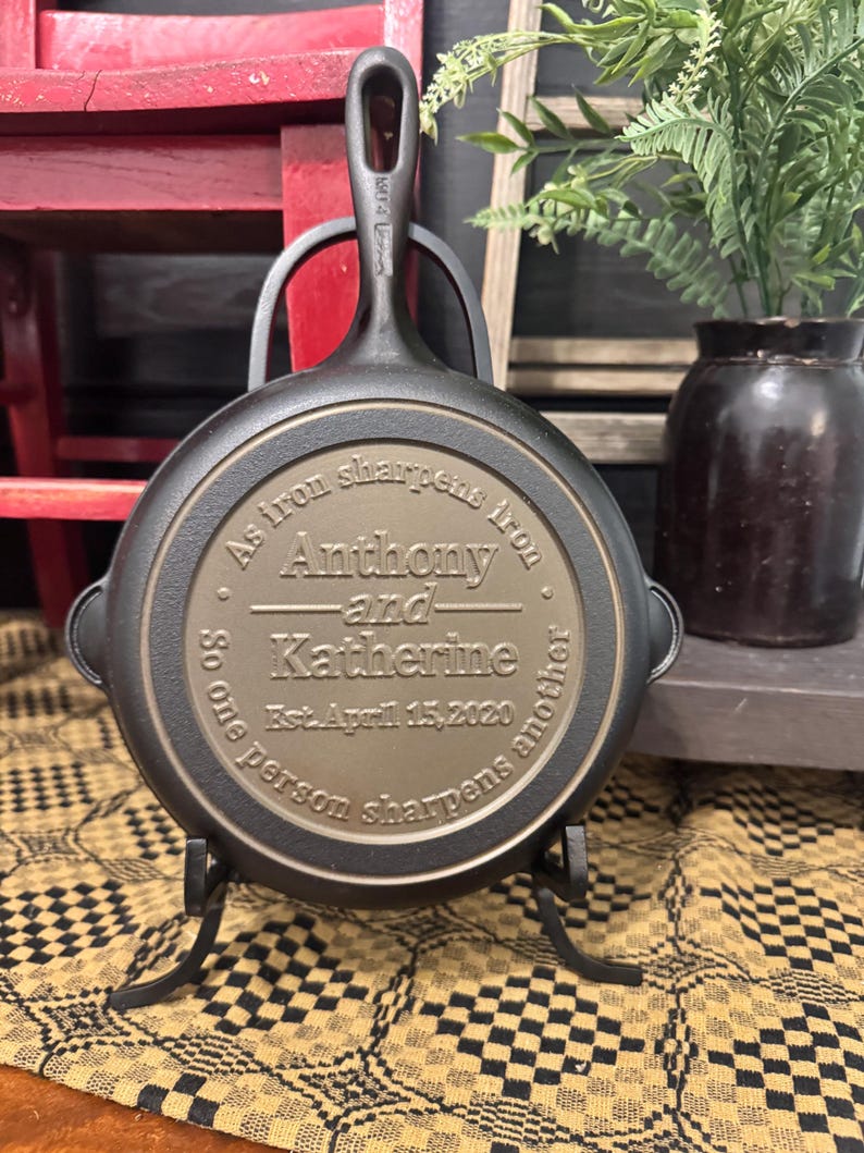 May include: A cast iron skillet with a handle, displayed on a black metal stand. The skillet's surface is engraved with the names "Anthony and Katherine" and the date "April 15, 2020", along with the phrase "As iron sharpens iron, so one person sharpens another."