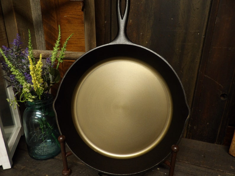 May include: A black cast iron skillet with a gold-colored interior. The skillet is on a metal stand.