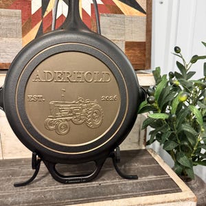 May include: A black cast iron skillet with a bronze-colored center. The center features the name "ADERHOLD" and a tractor illustration, with "EST. 2026" below. The skillet is displayed on a black metal stand.