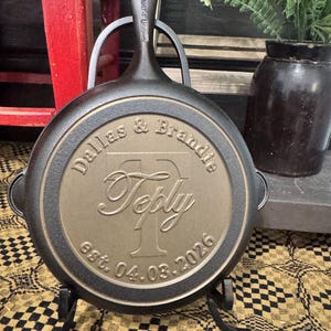 May include: A black cast iron skillet with a handle, resting on a black metal stand. The skillet's surface features the engraved names "Dallas & Brandie" above the name "Teply" and the date "est. 04.03.2026".