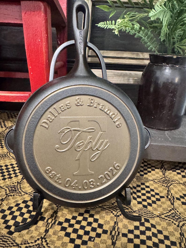 May include: A black cast iron skillet with a handle, resting on a black metal stand. The skillet's surface features the engraved names "Dallas & Brandie" above the name "Teply" and the date "est. 04.03.2026".