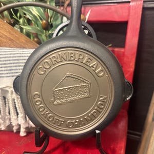 May include: A cast iron skillet with the words "CORNBREAD COOKOFF CHAMPION" embossed on the bottom. The skillet is black with a bronze-colored center, and a handle extends upwards. A slice of cornbread is depicted in the center.